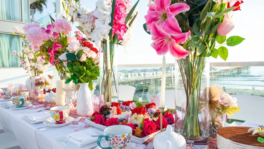 Decorated outdoor table with vibrant flowers, teacups, overlooking ocean and pier.