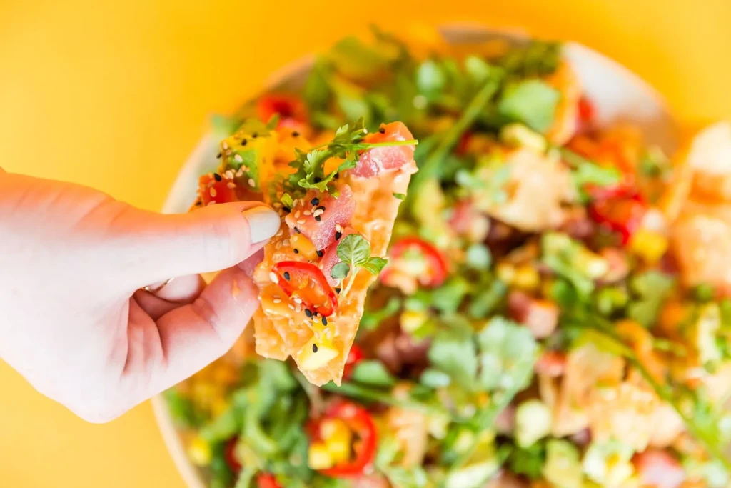 Hand holding a crisp cracker with colorful diced fish, fruit, and herbs.