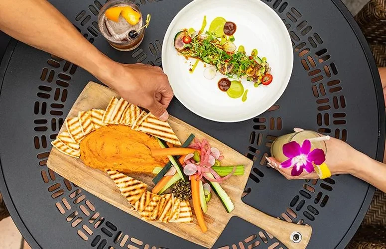 Overhead: Appetizers, plated dish, cocktails, and hands on a patterned table.