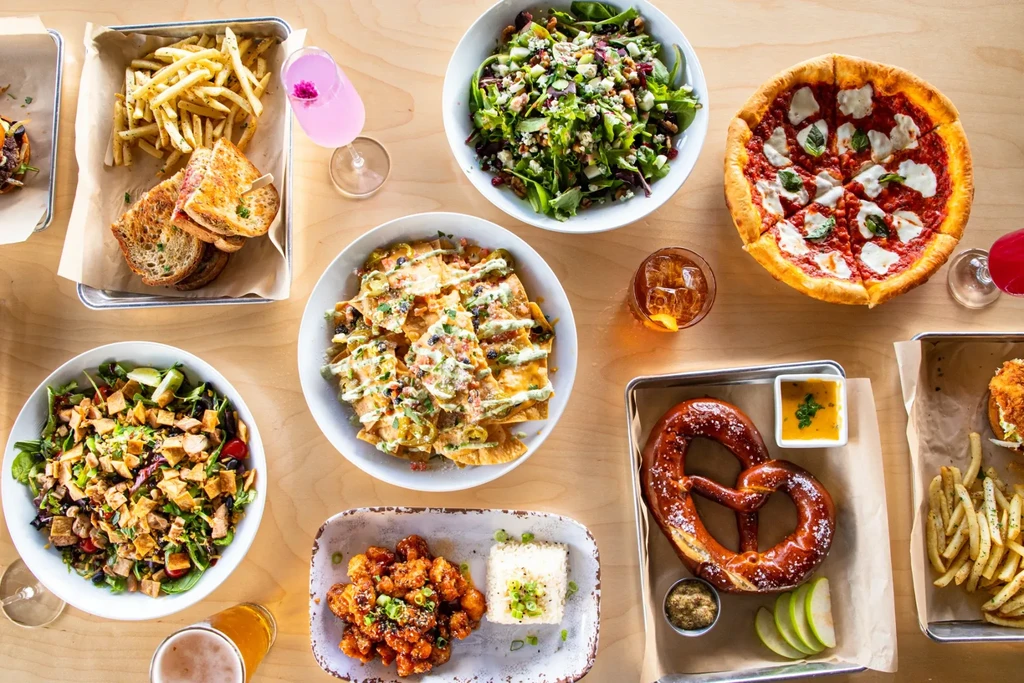 Diverse restaurant food spread: pizza, nachos, salads, pretzel, grilled cheese, drinks.