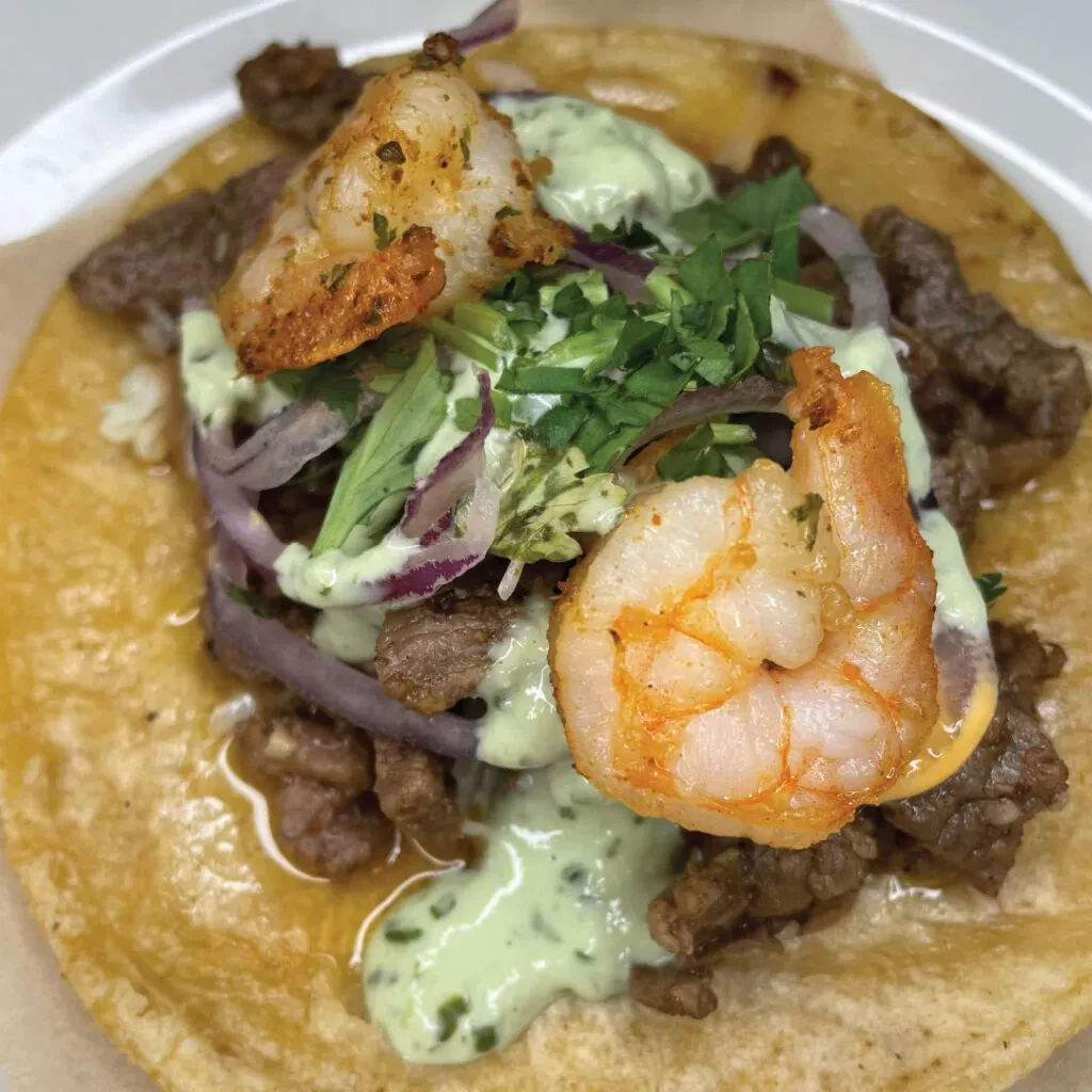 Taco with meat, two shrimp, green sauce, red onion, and cilantro.