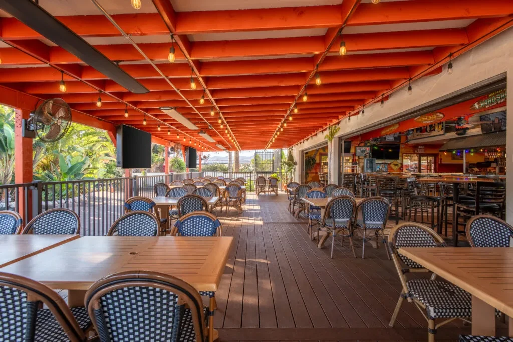 Empty restaurant patio with wood tables, tropical plants, and orange string-lit ceiling.