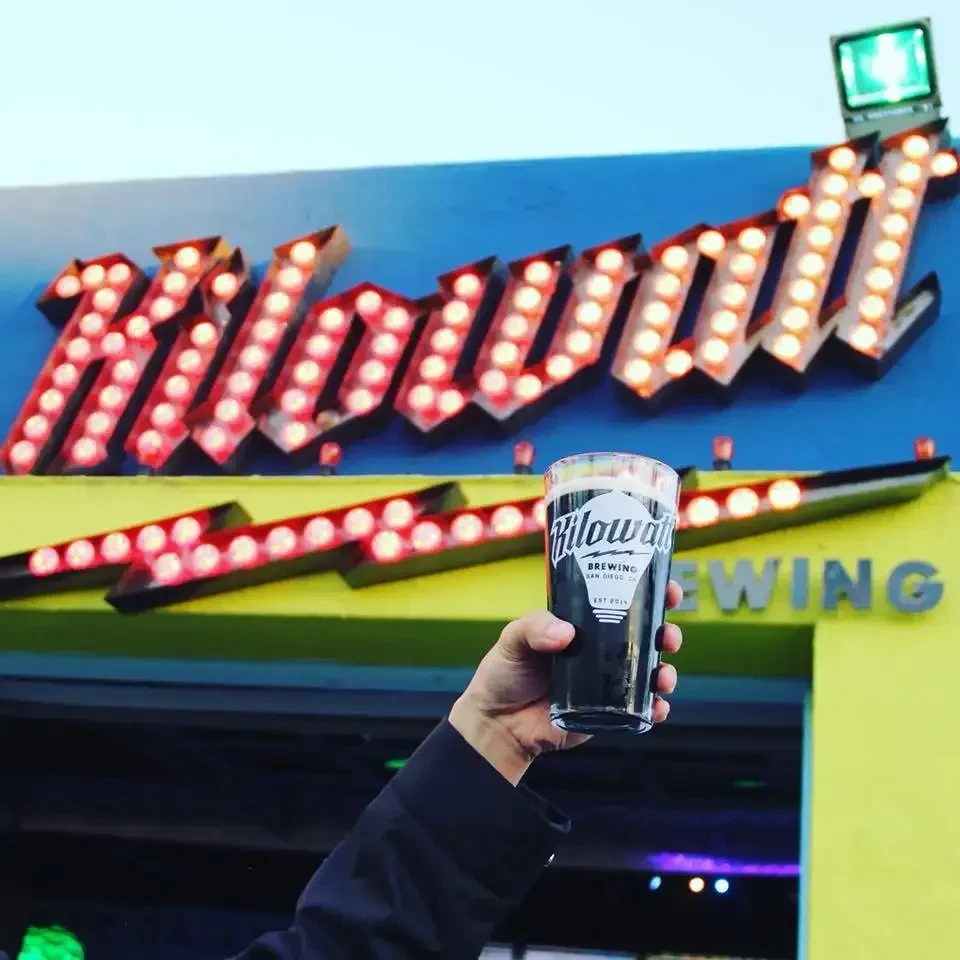 Hand holding beer glass against Kilowatt Brewing's vibrant illuminated sign.