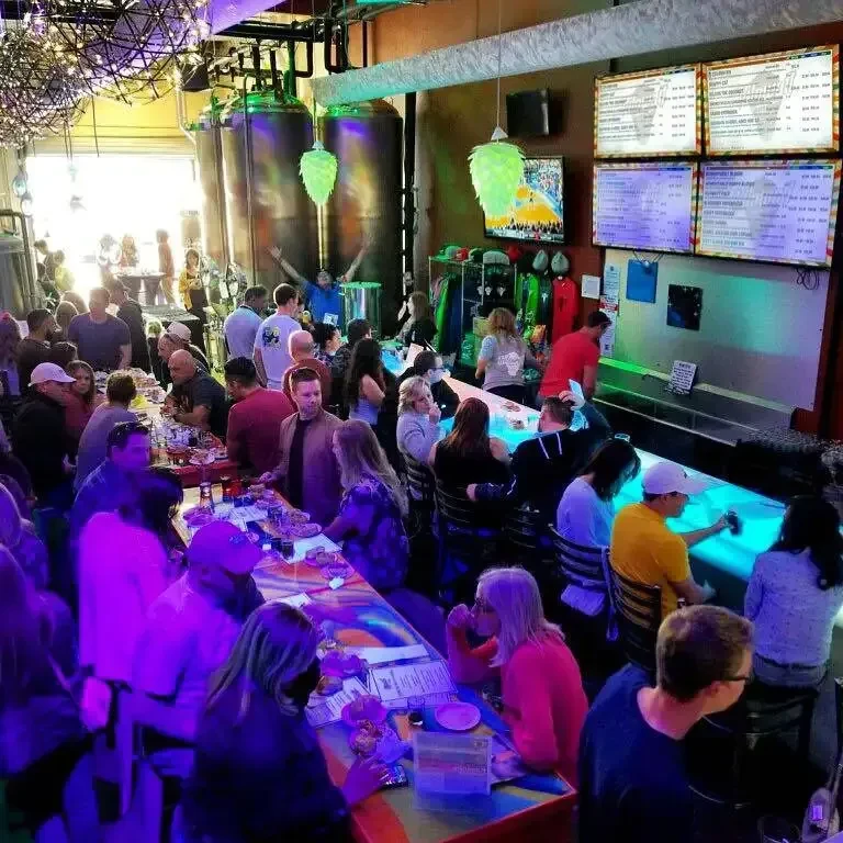 Busy brewery interior with fermentation tanks, people, and hop-shaped lights.