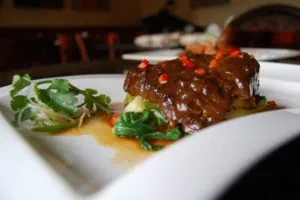 Plated braised meat with dark sauce, green vegetables, and red garnish.