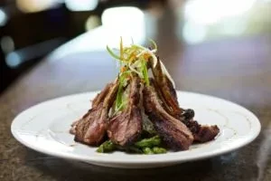 Stacked grilled lamb chops, green vegetables, and herbs on a white plate.