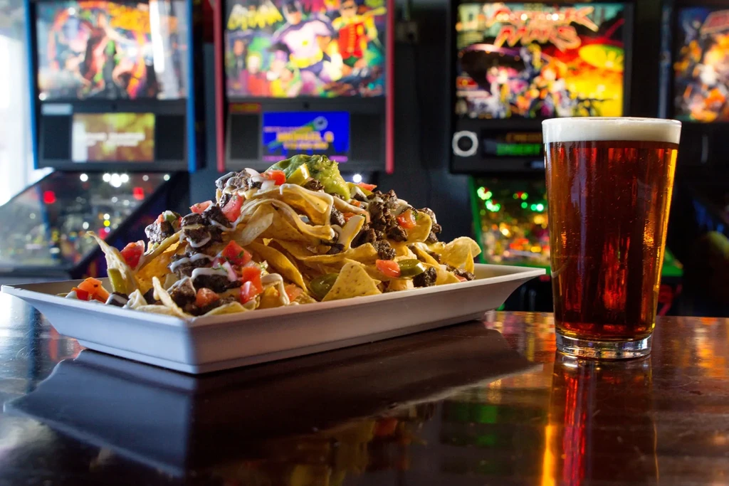 Loaded nachos and a beer in front of pinball machines.