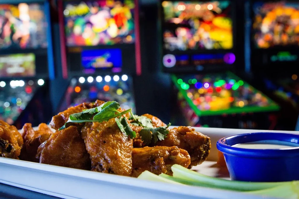 Plate of crispy, sauced chicken wings, celery, dip, blurred arcade.