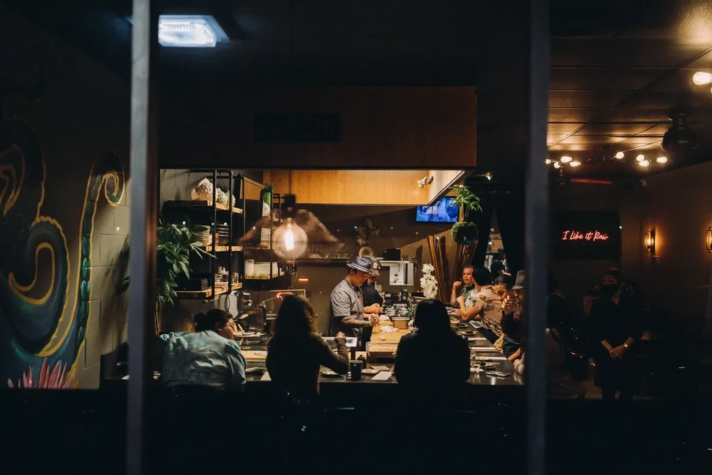 Dimly lit bar: chef, customers at counter, mural, neon sign.