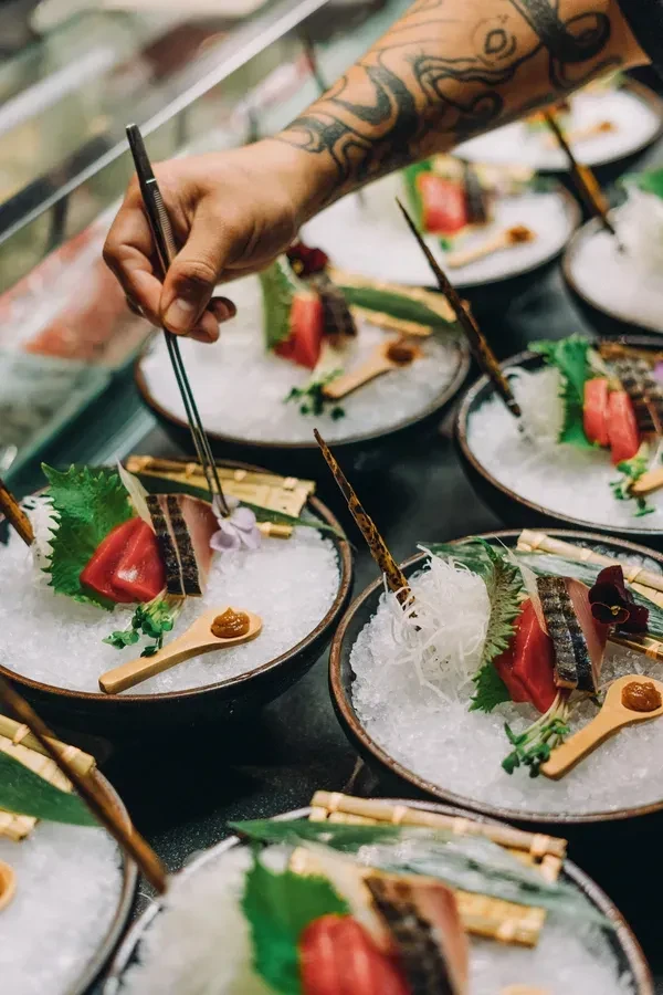 Tattooed chef's hand garnishes intricately plated sushi and sashimi dishes.
