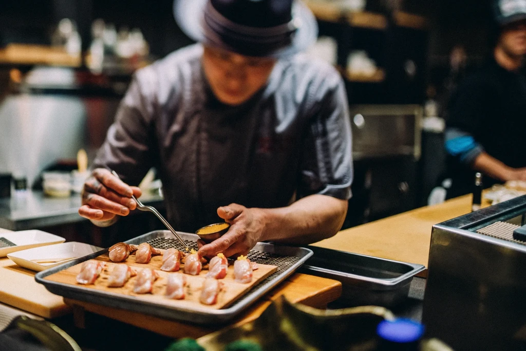 Chef uses tongs to add yellow topping to sushi on board.