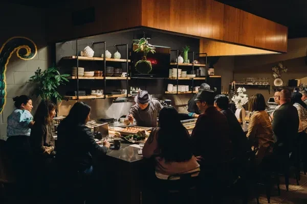 Chef prepares food for patrons at a dimly lit restaurant counter.