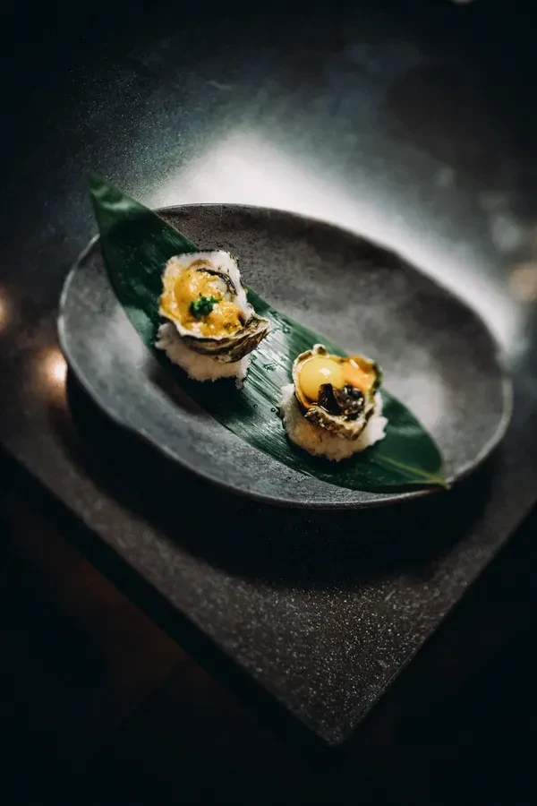 Two garnished oysters on rice, served on a green leaf and dark plate.