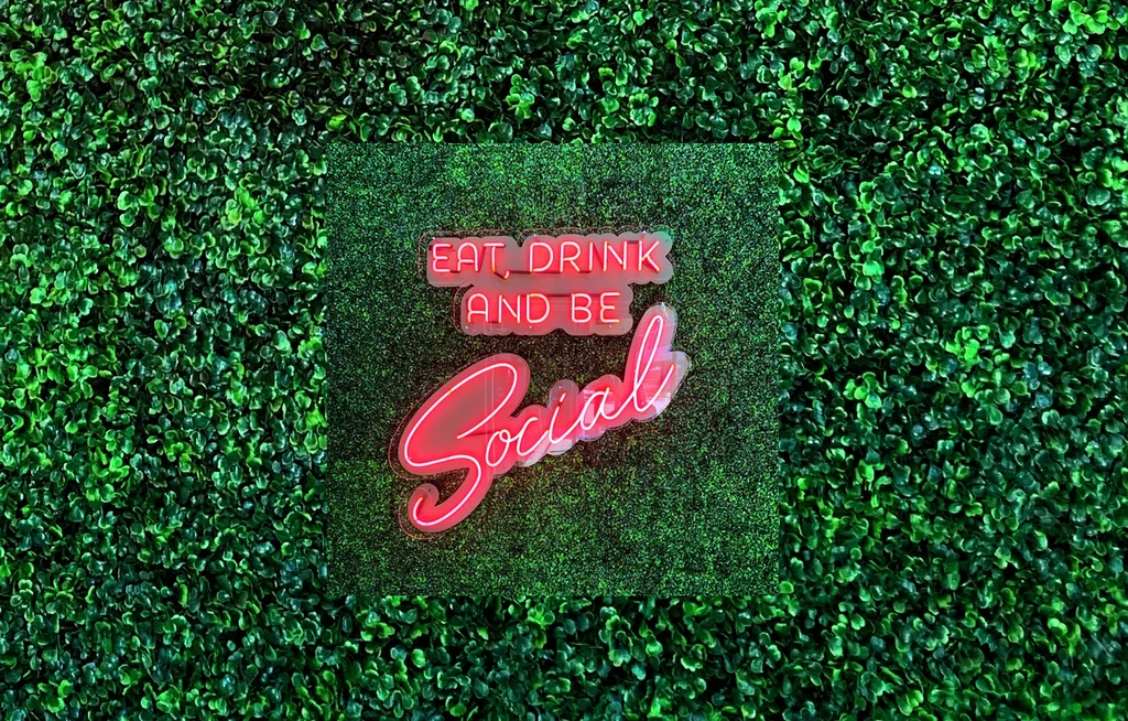 Red neon sign "EAT, DRINK AND BE Social" on a green foliage wall.