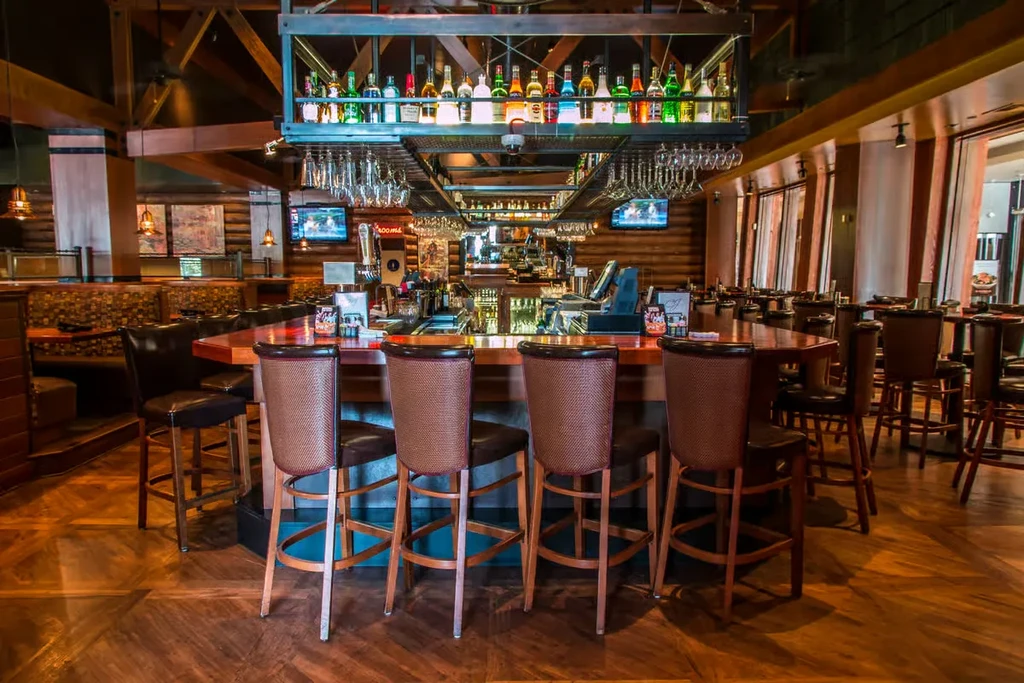 Empty, rustic modern bar interior with U-shaped wooden bar, illuminated bottles.