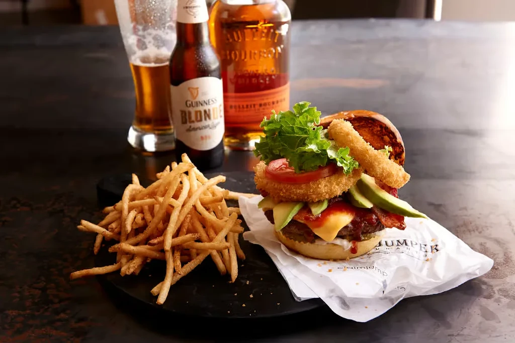 Elaborate burger, fries, beer, and bourbon whiskey on a dark surface.