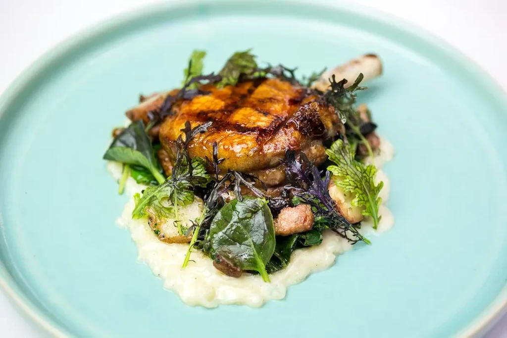 Seared bone-in meat, puree, greens, and potatoes on teal plate.