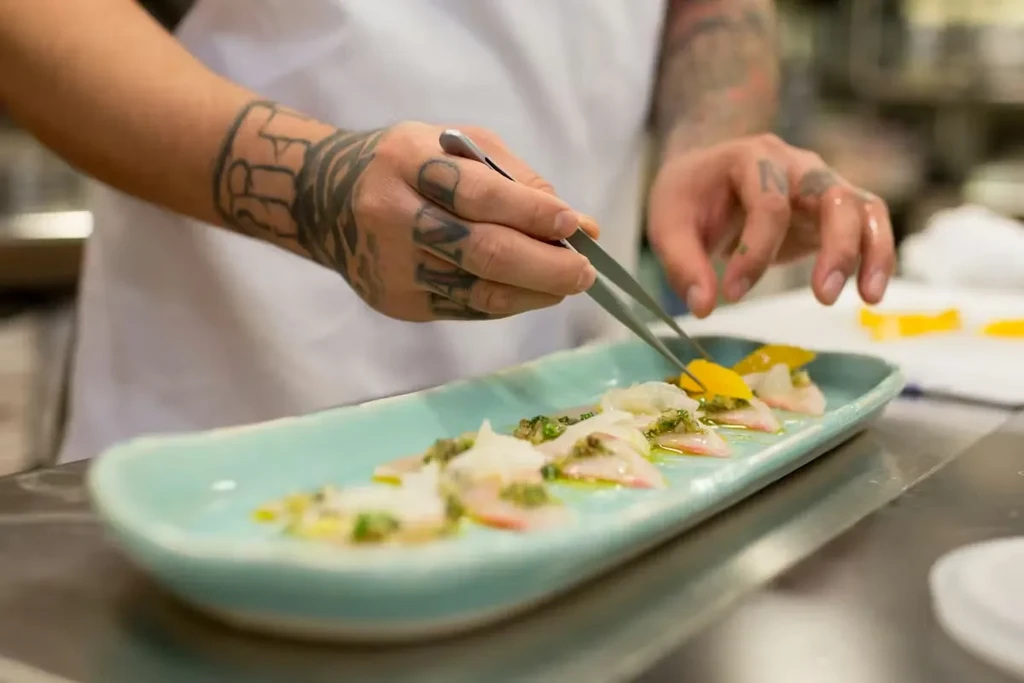 Tattooed chef's hands plating delicate crudo with citrus and green sauce.