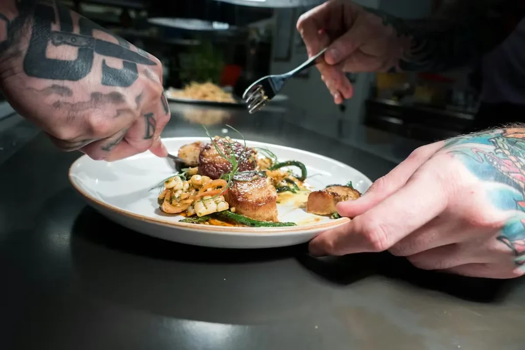 Tattooed hands meticulously adjust a plate of seared scallops and vegetables.