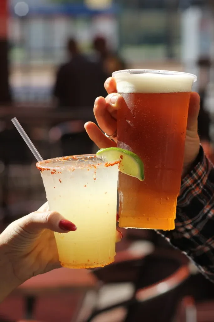 Two hands toast a chili-salted margarita and a beer with lime.