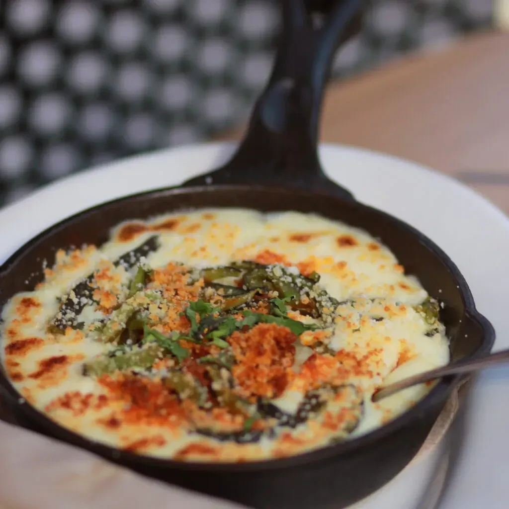 Bubbly cheesy gratin with green vegetables and crumbs in black skillet.