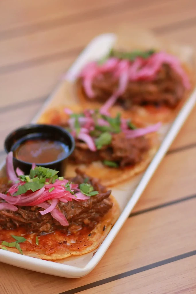 Three tacos with shredded meat, pickled onions, cilantro, and dipping sauce.