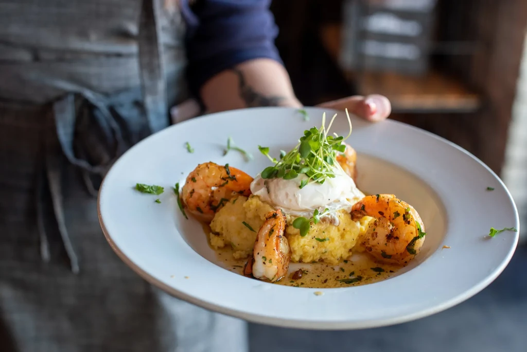 Close-up: server holds plated shrimp and grits with sauce and greens.