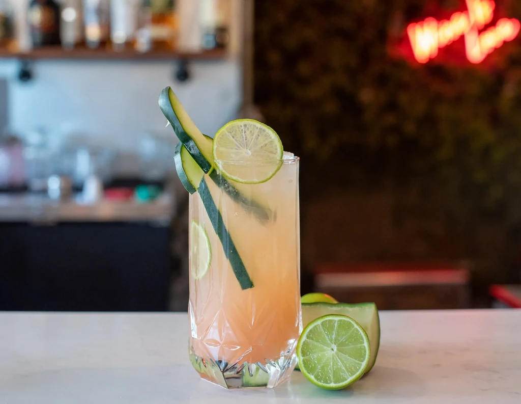 Faceted glass of amber cocktail garnished with cucumber and lime.