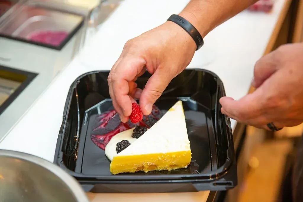 Hands garnish a triangular cheesecake slice with berries and cream.