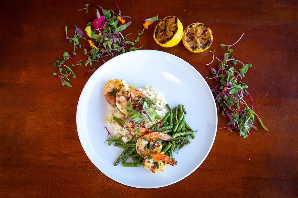 Plated grilled shrimp, creamy risotto, green beans, microgreens, and lemon.