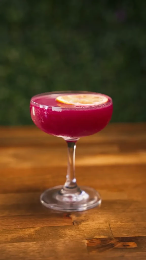 Vibrant magenta cocktail in coupe glass on wood with foliage.
