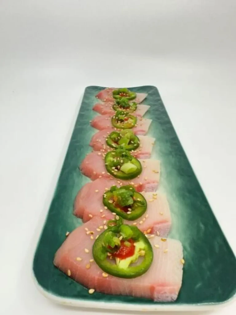 Raw fish slices garnished with jalapeño, sesame, and sauce on a teal plate.