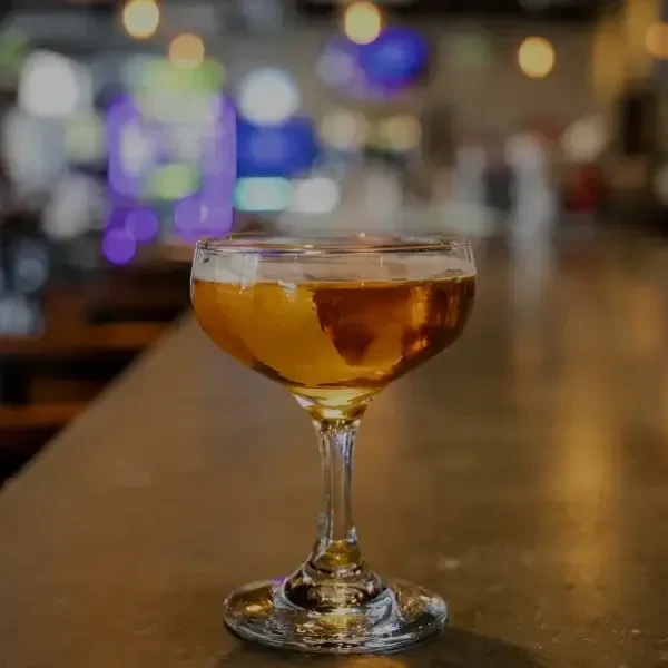 Amber cocktail in coupe glass on bar, blurred lights background.