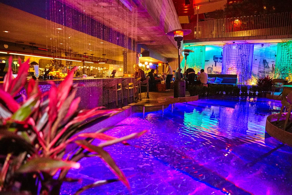 Vibrant bar interior with colorful mood lighting, patrons, and a glowing pool.