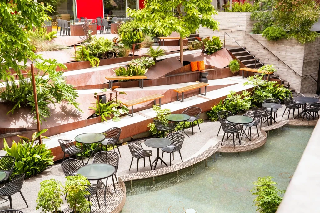 Modern multi-tiered outdoor lounge with rust planters, abundant greenery, and water.