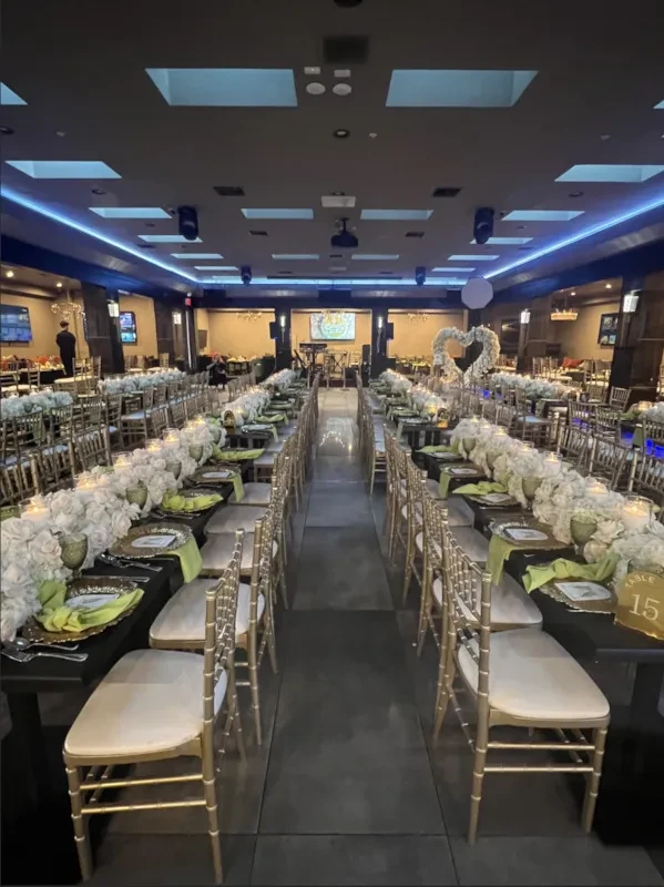 Elegant banquet hall with long tables, white flowers, gold chairs, blue lights.