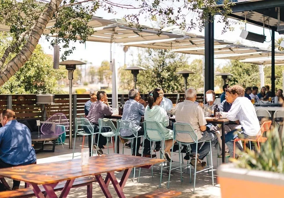 Sunny outdoor dining patio with people, large tree, canopy, and heaters.
