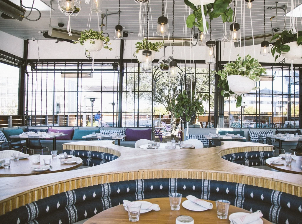 Modern restaurant interior with curved booth, colorful cushions, hanging lights, and plants.