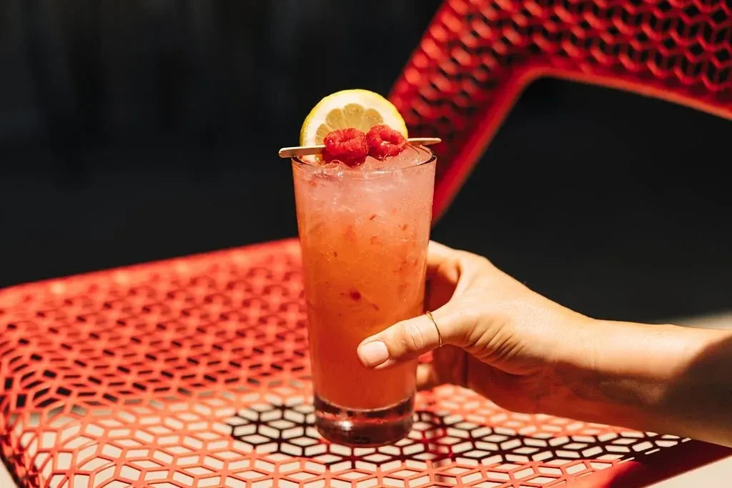Hand holding a garnished reddish-pink drink on a red hexagonal table.