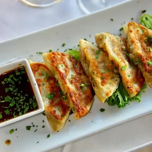 Six golden-brown pan-fried dumplings on white plate with herbs and sauce.