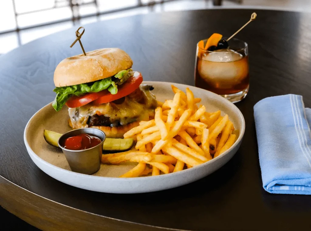 Burger, fries, ketchup, pickles, and Old Fashioned cocktail on dark table.