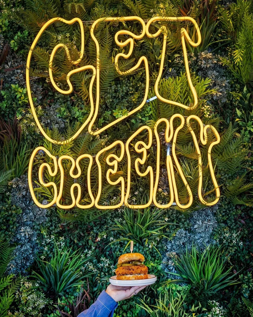 Stacked burger, 'GET CHEEKY' neon sign, and green plant wall.