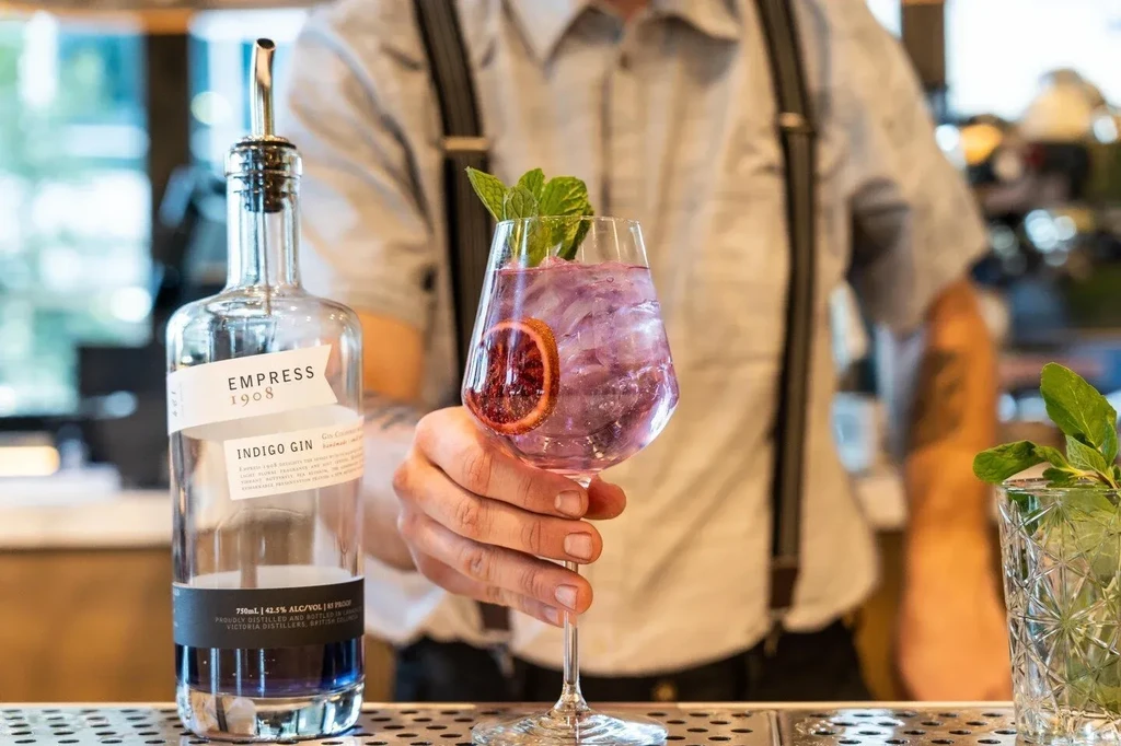 Bartender's hand holds purple cocktail next to Empress Indigo Gin bottle.