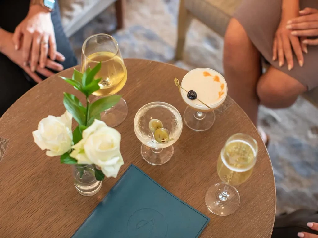 Round wooden table with four alcoholic drinks, white roses, and a menu.