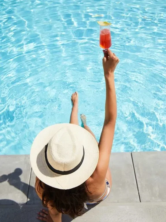 Overhead: Woman in straw hat holds drink, feet in blue pool water.