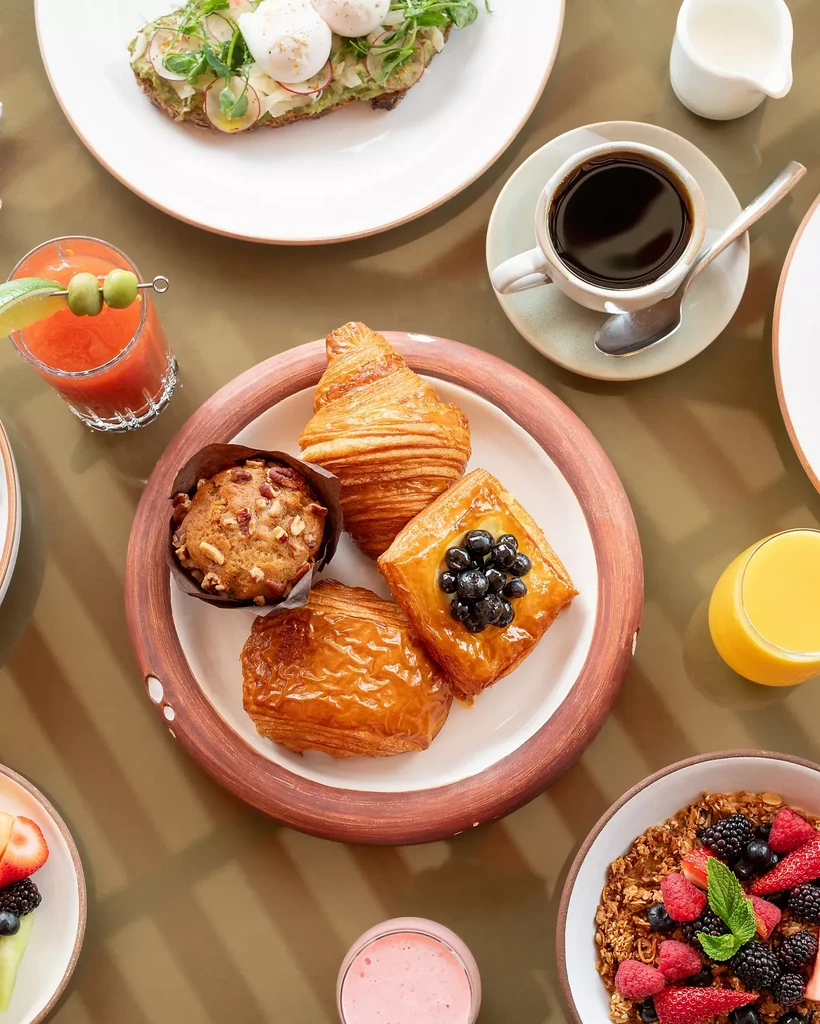 Flat-lay brunch spread with avocado toast, pastries, granola, and drinks.