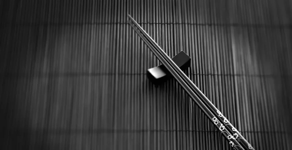Monochrome photo of dark patterned chopsticks on a rest and bamboo mat.