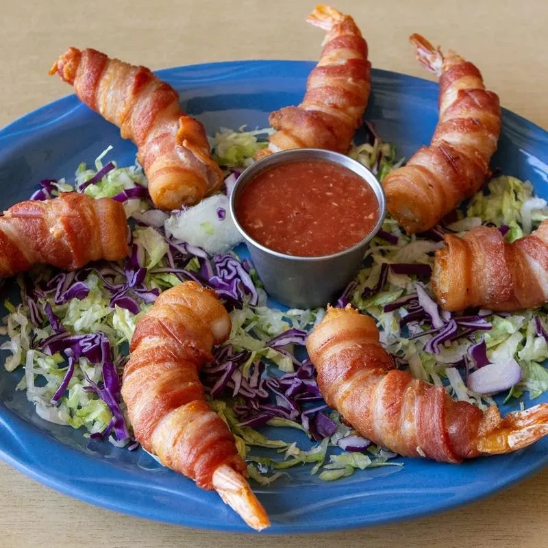 Six bacon-wrapped shrimp, dipping sauce, and shredded lettuce on blue plate.