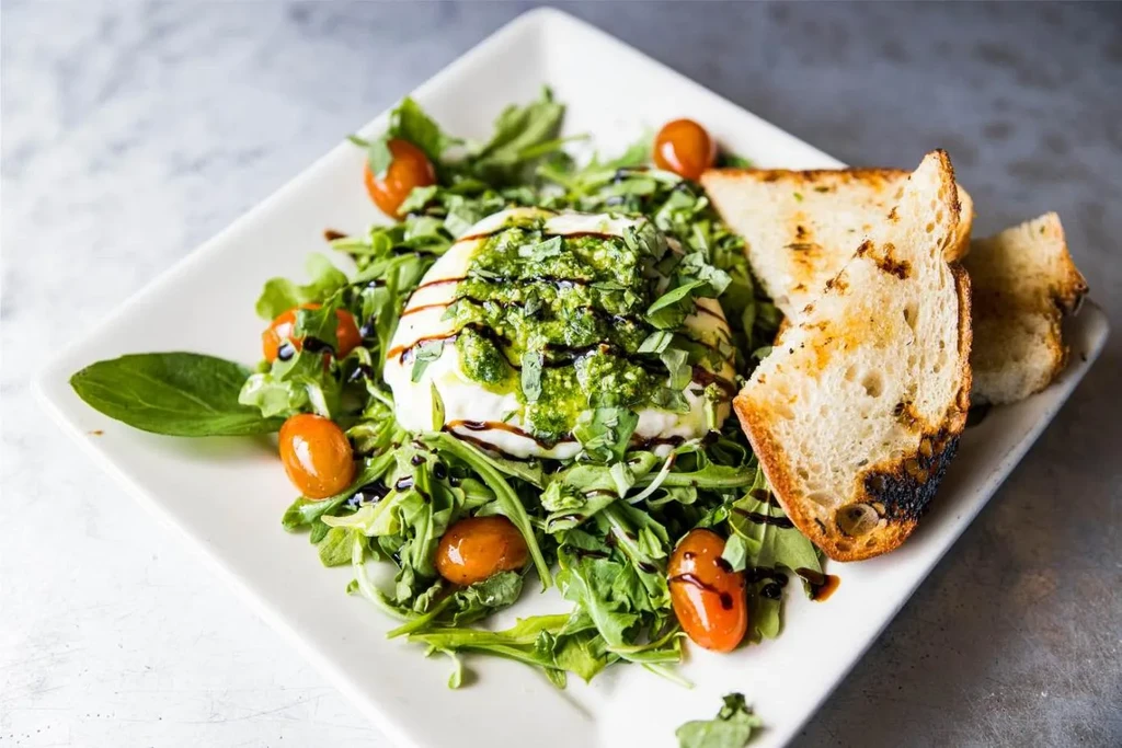 Plated burrata with pesto, arugula, cherry tomatoes, and toasted bread.