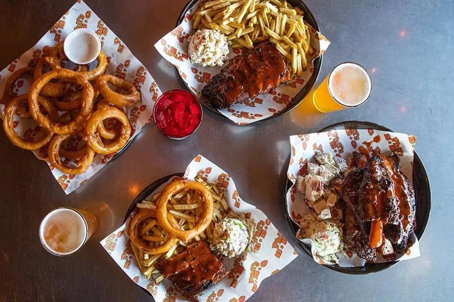 Barbecue meat platters, onion rings, fries, and beer on a table.